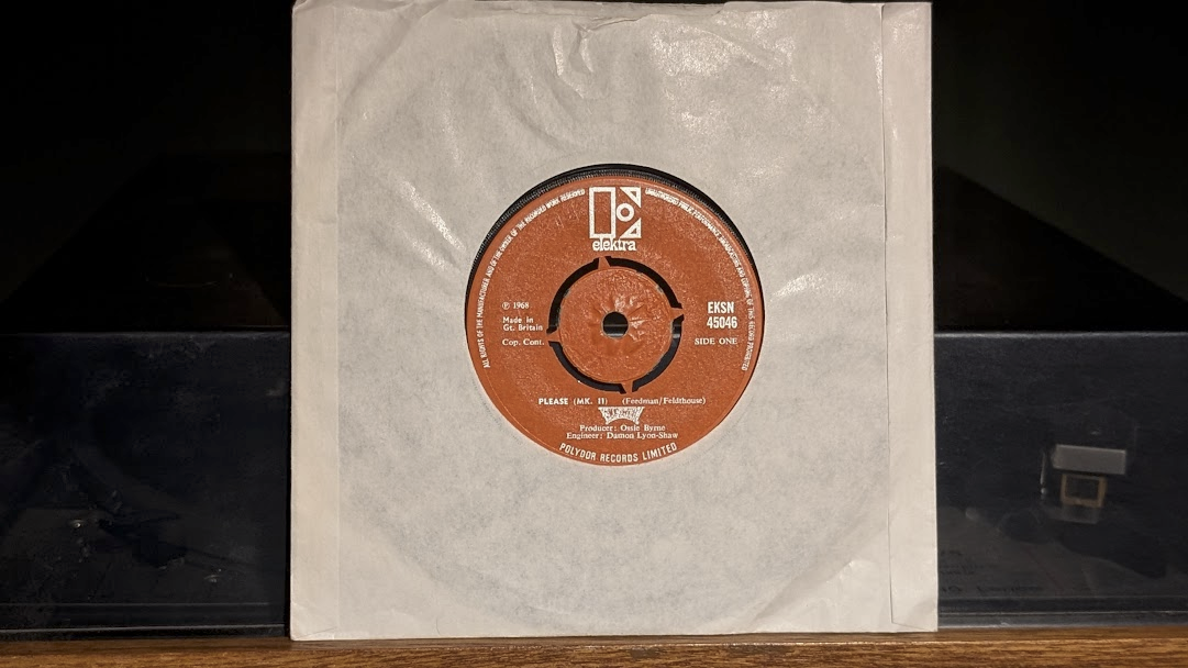 A copy of Eclection's "Please (Mk. II)" leaning against the smoked perspex cover of an old turntable.